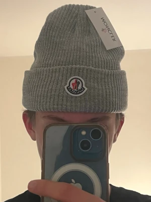 Grå Moncler mössa  - Snygg grå mössa från Moncler i ribbstickad ull med klassisk uppvikt kant och Moncler-logga framtill. Perfekt accessoar för kyliga dagar och ger en clean look till din outfit. Mössan har en neutral grå färg och är märkt med tag.