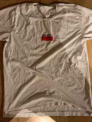 Vit HUGO BOSS t-shirt med tryck - Snygg vit t-shirt från HUGO BOSS med klassiskt tryck på bröstet i svart och rött. Modellen har rund hals och korta ärmar, perfekt för en chill och stilren look. Materialet är mjuk bomull som känns skönt mot huden. Kan gå ner i pris