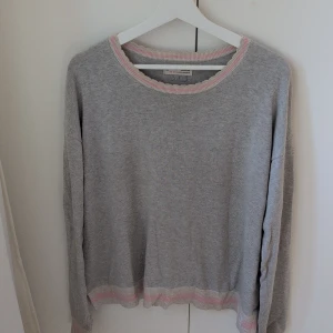 Grå stickad tröja Odd Molly - Säljer en grå stickad tröja från Odd Molly med ljusrosa och beige ribbade detaljer vid halsringning och nederkant. Tröjan har lång ärm och avslappnad passform, perfekt för chill dagar. Märkeslapp på nederkanten och broderad logga på ärmen.