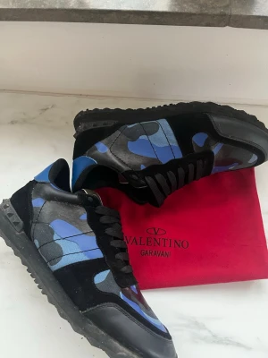 Valentino Rockrunner blå sneakers - Säljer ett par Valentino Rockrunner sneakers med blått och svart camouflage-mönster. Skorna har svarta snören, platt sula och detaljer i både mocka och skinn. Metallplatta med Valentino Garavani-logga på plösen. Riktigt snygga och unika sneakers för dig som gillar statement pieces.