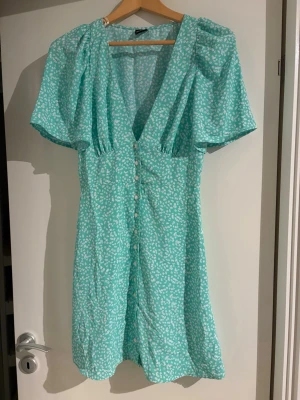 Mintgrön blommig klänning Gina Tricot - Superfin mintgrön kortklänning från Gina Tricot med vita småblommor, v-ringning och knappar framtill. Klänningen har korta, puffiga ärmar och en lätt, flowy passform. Perfekt för varma dagar och somriga vibes!