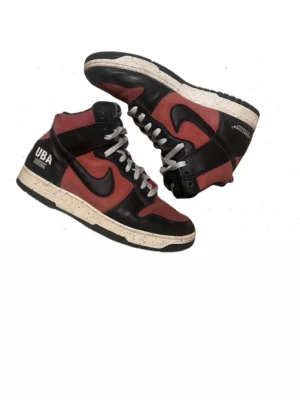 Nike Dunk High UBA x Undercover  sneakers - Säljer ett par Nike Dunk High UBA x Undercover sneakers i svart och röd med vita snören och beige sula med svarta prickar. Skorna har högt skaft, rund tå och är tillverkade i skinn. UBA- och Undercover-logga på hälen. Perfekt för dig som gillar unika collabs.