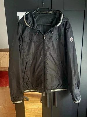 Svart vindjacka från Moncler - Snygg svart vindjacka från Moncler med huva och dragkedja framtill. Jackan har två bröstfickor med knappar och ribbade muddar med vita ränder vid ärmslut och nederkant. Perfekt för dig som gillar stilren och sportig look. Bra skick skulle ja säga
