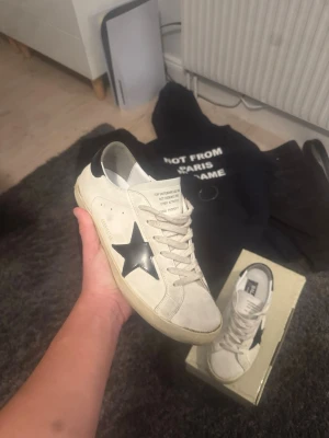 Golden Goose Superstar sneakers vit/svart - Golden Goose Superstar sneakers i vit mocka med svart stjärndetalj på sidan och svart häl. Klassisk rund tå, snörning och platt sula. Skorna har en cool vintage vibe och levereras med originalkartong och dustbag. Perfekta för dig som gillar streetstyle och unika detaljer.