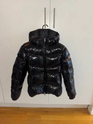 Svart pufferjacka från Colmar - Säljer en svart, glansig pufferjacka från Colmar med huva och dragkedja framtill. Jackan har ett stilrent, quiltat mönster och logotyp på ärmen. Perfekt för dig som vill ha en varm och snygg jacka med modern vibe.