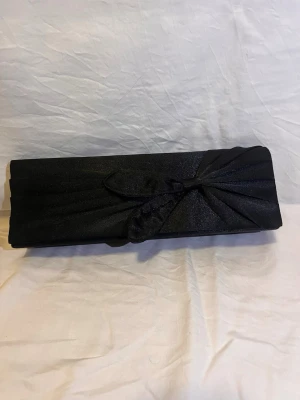 Kuvertväska - Elegant svart kuvertväska i satin med en dekorativ rosett framtill. Väskan har en avlång och stilren form, perfekt för att bära det viktigaste. Materialet har en lätt glansig finish som ger en lyxig känsla. Skedjan är borttappad.