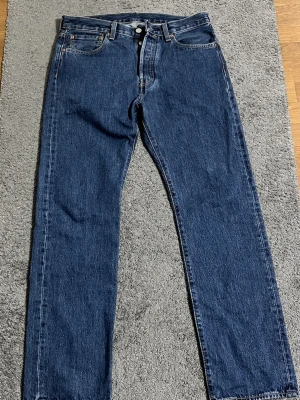 Levi's 501 jeans - Klassiska Levi's 501 jeans i mörkblå denim med raka ben och normal passform. Byxorna har fem fickor, knappgylf och tydliga kontrastsömmar. Ikonisk läderpatch bak i midjan och röd Levi's-tagg på bakfickan. Perfekta för en avslappnad stil.