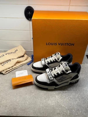 Louis Vuitton svartvita sneakers - Louis Vuitton sneakers i svart och vitt med klassiska LV-monogramdetaljer och broderad logga på sidan. Skorna har snörning, meshpaneler och en dekorativ rem med ikoniska symboler. Platt sula och rund tå, tillverkade i skinn och syntetmaterial.