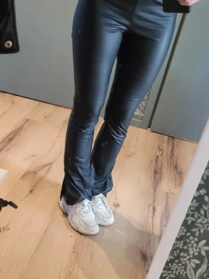 Svarta bootcut byxor från Shein XS - Säljer ett par svarta bootcut byxor från Shein i storlek XS. Byxorna har en glansig finish, elastisk midja och är tillverkade i syntetmaterial. De har slits vid bensluten och sitter tajt upptill med utsvängda ben. Perfekt för dig som gillar trendiga och bekväma byxor. Aldrig använda.