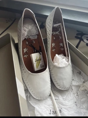 damskor  - Snygga vita ballerinaskor från Jana med dekorativa hål längs sidorna och blommigt mönster på innersulan. Skorna har rund tå, platt sula och är tillverkade i mjukt skinn för extra komfort. Perfekta för dig som vill ha en stilren och bekväm sko.