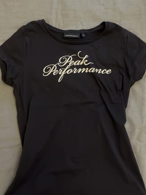 Mörk blå Peak Performance t-shirt - Svart t-shirt från Peak Performance med vit logga tryckt på bröstet. Klassisk rund halsringning och korta ärmar. Perfekt för en sportig och stilren look.