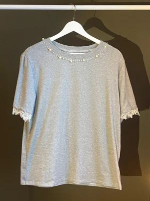 Grå topp med pärlor och spetsdetaljer - Snygg grå topp med pärlor runt halsringningen och spetsdetaljer vid ärmsluten. T-shirten har en rak passform och korta ärmar. Perfekt för dig som gillar enkla men ändå unika detaljer. Materialet känns mjukt och bekvämt. Köpt från SHEIN, aldrig använt🩶