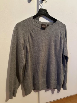 Grå stickad tröja från Oscar Jacobson - Klassisk grå stickad tröja från Oscar Jacobson med rund halsringning och ribbade muddar. Tillverkad i Italien och har en enkel, stilren design som passar till det mesta. Strl S.