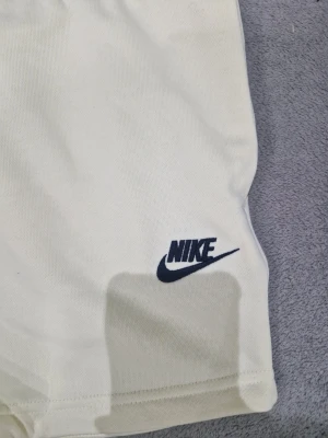 Vita Nike shorts med snörning - Snygga vita shorts från Nike med klassisk logga broderad i svart på benet. Shortsen har snörning i midjan för perfekt passform och är tillverkade i mjuk bomull, vilket gör dem riktigt sköna att bära. Perfekta för en sportig och avslappnad stil.