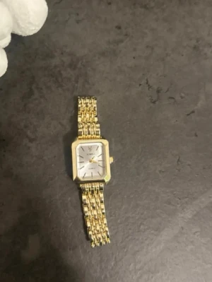 Guldfärgad fyrkantig klocka från ximili  - Stilren guldfärgad klocka från Komono med fyrkantig urtavla och länkarmband i metall. Urtavlan är vit med guldiga visare och index, vilket ger en klassisk och elegant look. Perfekt accessoar för dig som gillar tidlös design.