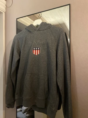 Grå hoodie från GANT  - Säljer en grå hoodie från GANT med broderad logga och amerikanskt sköldmärke på bröstet. Hoodien har känguruficka framtill och en klassisk huva. Perfekt för en chill och avslappnad stil.