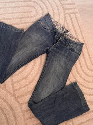 Blå bootcut jeans med blommigt foder - Snygga blå bootcut jeans med blommigt innerfoder i midjan. Klassisk femficksmodell och normal passform. Jeansen har en lätt tvättad look och är perfekta för dig som gillar en avslappnad stil. 