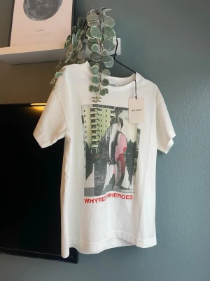 Helt ny Vit t-shirt med tryck från Whyred - Säljer en vit t-shirt från Whyred i storlek S med coolt grafiskt tryck framtill och texten 'WHYRED FOR HEROES' i rött. Lite oversized passform med korta ärmar och rund hals. Perfekt för dig som gillar streetstyle och vill sticka ut lite extra. Helt ny med prislappar.