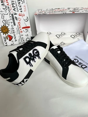 Dolce & Gabbana vita och svarta sneakers - Snygga vita sneakers från Dolce & Gabbana med svarta detaljer och stort D&G-tryck i graffiti-stil på sidan. Skorna har snörning, rund tå och är tillverkade i skinn med kontrasterande svarta partier och logotypdetaljer på hälen.
