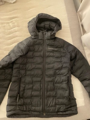 Svart dunjacka Peak Performance - Snygg svart dunjacka från Peak Performance med huva och dragkedja framtill. Jackan har två sidofickor (ena fickan har ett hål, inget som syns på utisidan)  med dragkedja och diskret logga på bröstet. Perfekt för kalla dagar och enkel att matcha med allt.