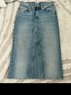 Blå jeanskjol från 157 - Rak, blå lång jeanskjol från 157 med klassiska fem fickor och knappgylf. Kjolen har en slits framtill och är gjord i slitstarkt denimtyg. Perfekt för dig som gillar en enkel och tidlös stil.