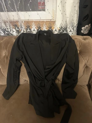 Svart omlottkavaj med bälte - Stilren svart kavaj med omlottdesign och matchande bälte i midjan. Kavajen har långa ärmar och klassisk krage, perfekt för en trendig och clean look. Materialet känns mjukt och följsamt, vilket ger en snygg passform.