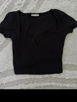 Svart ribbad croppad topp från Zara - Säljer en svart croppad topp från Zara med ribbad struktur och snygg v-ringad urringning. Toppen har korta ärmar och sitter tight, perfekt till jeans eller kjol. Enkel men trendig design som passar till många olika looks.