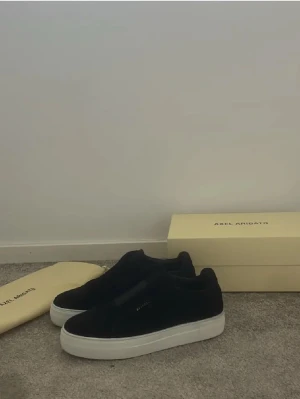 Svarta sneakers från Axel Arigato - Stilrena svarta sneakers från Axel Arigato med ovandel i mocka och vit platt sula. Diskret logga i guld på sidan och minimalistisk design utan snörning. Perfekta för dig som gillar en clean och modern look.