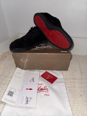 Svarta Louboutin sneakers med nitar - Säljer ett par svarta sneakers från Christian Louboutin med ikoniska röda sulor och coola svarta nitar över hela tån. Skorna är i mocka och har snörning. Perfekta för dig som vill sticka ut med en lyxig och edgy stil.