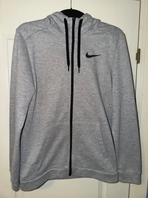 Grå zip hoodie från Nike - Snygg grå zip hoodie från Nike med svart logga på bröstet och svarta dragsnören i huvan. Tillverkad i mjukt sweatshirtmaterial och har två stora fickor framtill. Perfekt för en avslappnad och sportig stil.