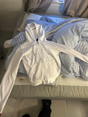 Vit zip hoodie från Juicy Couture - Säljer en vit hoodie från Juicy Couture med dragkedja framtill, stor huva och fickor på magen. Hoodien har snören i huvan och ett diskret Juicy Couture-logotyp broderat på bröstet. Perfekt för en chill och stilren look.