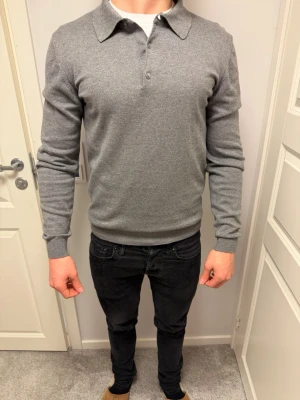 Grå pikéstickad tröja med krage - Snygg grå långärmad tröja med klassisk pikékrage och knappar framtill. Tröjan har ribbade muddar vid ärmslut och nederkant, vilket ger en clean och stilren look. Perfekt till jeans eller chinos för en avslappnad men ändå uppklädd stil. Ts Ai