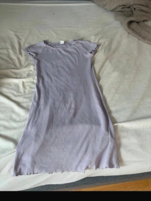 Ljuslila ribbad kortklänning - Säljer en ljuslila ribbad kortklänning med korta ärmar och vågig kant vid halsringning och ärmslut. Klänningen har en enkel, figurnära passform och är gjord i mjukt bomullsmaterial. Perfekt för en avslappnad och trendig look.