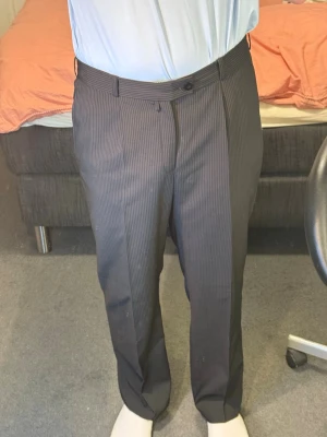 Mörkgrå randiga kostymbyxor - Snygga mörkgrå kostymbyxor med tunna vertikala ränder. Klassisk rak passform med bälteshällor och knappstängning i midjan. Perfekta för en stilren look. Materialet känns mjukt och har en lätt glans.