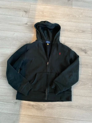Svart hoodie från Polo Ralph Lauren - Svart hoodie från Polo Ralph Lauren med dragkedja framtill och klassisk röd logga broderad på bröstet. Säljer då den inte passar mig längre.