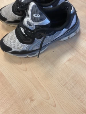 Svarta och vita sneakers från Asics - Säljer ett par snygga svarta och vita sneakers från Asics. Klassisk sportig design med snörning och kontrasterande detaljer. Perfekta för dig som gillar stilrena och bekväma skor till vardags. Sulan är vit med svarta inslag och ovandelen har Asics-loggan.