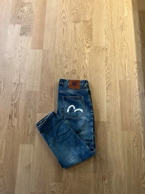Blå jeans från Evisu med målad ficka - Snygga blå jeans från Evisu med deras ikoniska vita målade logga på bakfickorna och läderpatch i midjan. Jeansen har rak passform och klassisk femficksdesign. Fint skick med slitningar som självklart är modellen