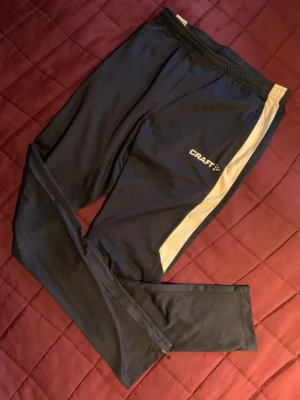 Marinblå träningsbyxor från Craft - Snygga svarta träningsbyxor från Craft med vit sidopanel och logga på låret. Byxorna har smal passform, resår i midjan och dragkedja vid benslut. Perfekta för löpning eller gymmet.