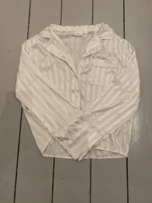 Vit randig satinpyjamas från Gina Tricot - Snygg vit pyjamaströja från Gina Tricot i glansigt satinmaterial med diskreta ränder. Klassisk krage, knappstängning framtill och en bröstficka. Perfekt för mysiga kvällar hemma eller när du vill känna dig extra chill.