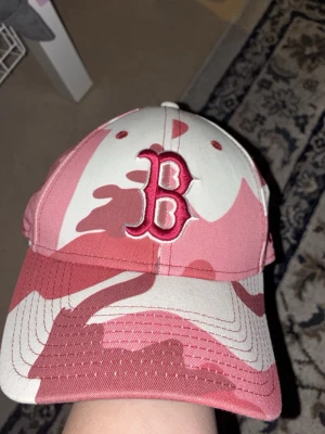 Rosa camo keps med B-logga - Säljer en riktigt snygg keps i rosa och vit camo med en stor broderad B-logga framtill. Justerbart spänne bak för perfekt passform. Perfekt accessoar för dig som vill sticka ut lite extra. Materialet känns som bomull.