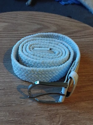 Vitt flätat bälte med silverfärgat spänne - Snyggt vitt flätat bälte med ett stilrent silverfärgat metallspänne. Bältet är gjort i ett mjukt syntetmaterial och har en klassisk design som passar till många olika outfits. Bältet är ungefär 107 cm långt.