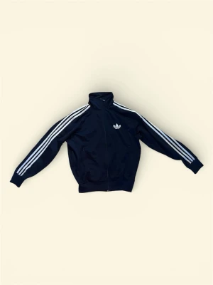 Mörkblå Adidas track jacket - Klassisk mörkblå Adidas track jacket med dragkedja, vita ränder längs ärmarna och Adidas logga på bröstet. Jackan har hög krage och ribbade muddar. Perfekt för en sportig och avslappnad stil.