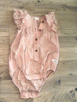 Rosa body med spetsärmar från Newbie - Supersöt rosa body från Newbie i storlek 62, med dekorativa spetsärmar och knappar framtill. Tillverkad i mjukt, luftigt tyg och har tryckknappar i grenen för enkel påklädning. Perfekt för små barn som vill vara både bekväma och stiliga.