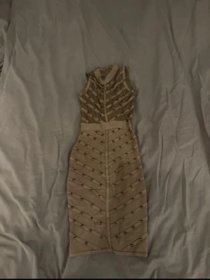 Beige ärmlös klänning med nitar - Snygg beige ärmlös klänning med markerad midja och dekorativa guldfärgade nitar i mönster över hela klänningen. Klänningen har en kroppsnära passform och dragkedja i ryggen. Perfekt för dig som vill sticka ut med en unik look.