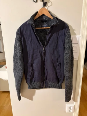 Woolrich cardigan - Snygg grå stickad cardigan från Woolrich med quiltad front och ärmar i grovstickat material. Tröjan har dragkedja, hög krage och två fickor framtill. Perfekt för lager-på-lager och chill höststil. Fits S/M