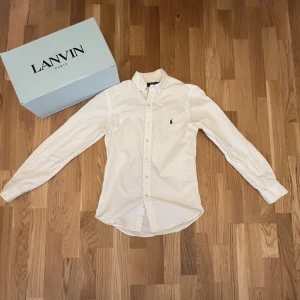 Vit skjorta från Ralph Lauren - Vit skjorta från Ralph Lauren med klassisk button-down krage och broderad logga på bröstet. Skjortan har lång ärm, knappar hela vägen och är tillverkad i mjuk bomull. Perfekt för dig som gillar stilrena och tidlösa plagg.