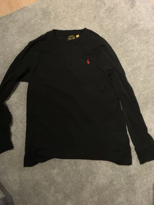 Svart långärmad topp i bomull - En stilren svart långärmad topp i mjuk bomull. Enkel design med rund halsringning och smal passform, perfekt att matcha med jeans eller kjol. Liten röd detalj vid halsen för en extra touch.