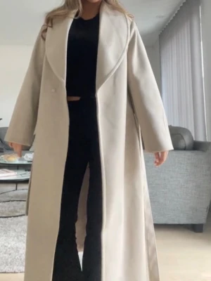 Beige lång kappa med bred krage - Säljer denna jättesnygga kappan då jag redan har en lång kappa. Den är i storlek L men funkar för M också om man vill ha lite oversized som jag på bilden. Den är helt oanvänd!