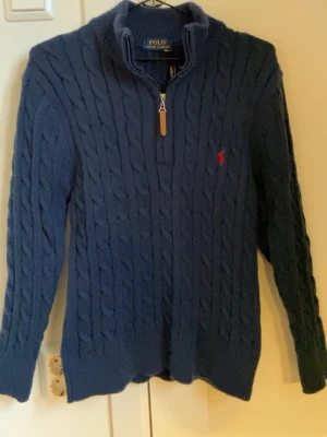 Polo Ralph Lauren stickad tröja - Mörkblå kabelstickad tröja från Polo Ralph Lauren med halv dragkedja och hög krage. Tröjan har det klassiska röda Polo-logot broderat på bröstet och ribbade muddar vid ärmslut och nederkant. Perfekt för en stilren och avslappnad look. Tröjan är oanvänd!
