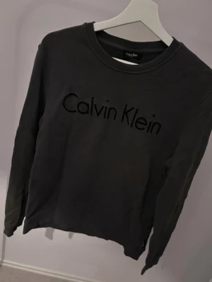 Svart sweatshirt från Calvin Klein - Snygg svart sweatshirt från Calvin Klein med broderad logga framtill. Tröjan har rund halsringning och långa ärmar. Perfekt för en clean och stilren look. Tillverkad i mjukt material för extra komfort.